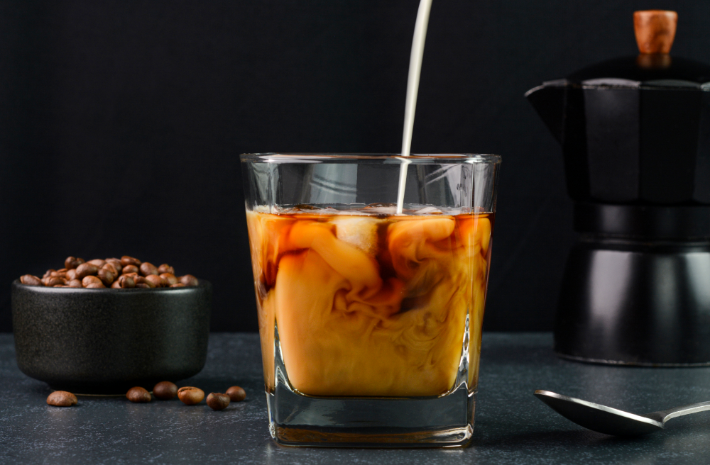 Creamer is added to a freshly made cold brew coffee serving in a glass with coffee beans and the coffee maker both in the background.