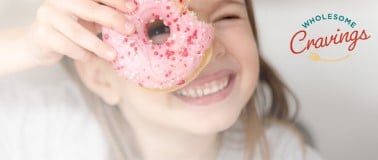 A young girl smiling and looking through the hole of a pink frosted donut